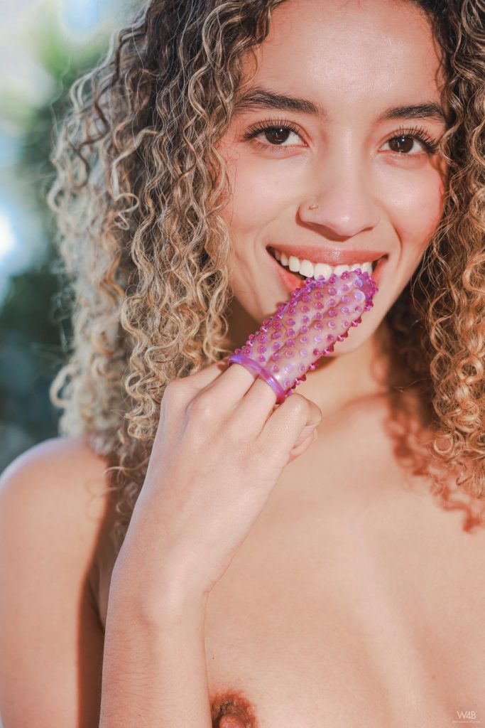 Close up of a nude girl with curly blonde hair smiles while holding up some kind of rubber finger sleeve to her face.