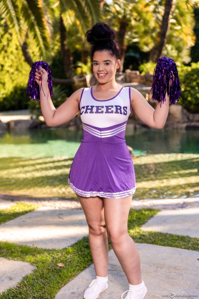 A young woman in a purple and white cheerleader outfit holds purple pom-poms and smiles while standing outdoors in front of a pool, with trees and greenery in the background.