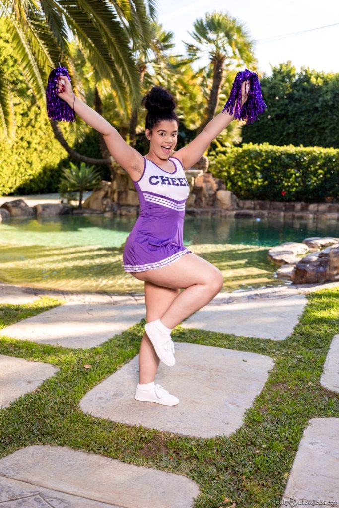 A cheerleader in a purple uniform with CHEER written on it poses outdoors on one leg, holding purple pom-poms up high, with a pool, palm trees, and greenery in the background.