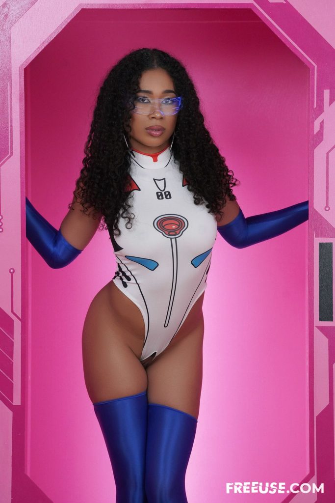 A woman with curly hair and glasses poses in a futuristic, form-fitting bodysuit and long blue gloves and stockings, standing in a pink doorway with a sci-fi theme.