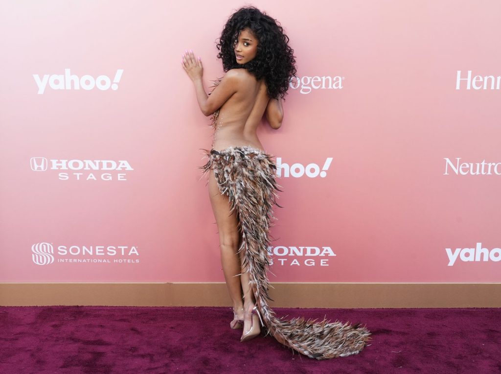 A woman with curly hair poses on a maroon carpet against a pink backdrop, wearing a nude-toned outfit adorned with feathers and a dramatic feathered train.
