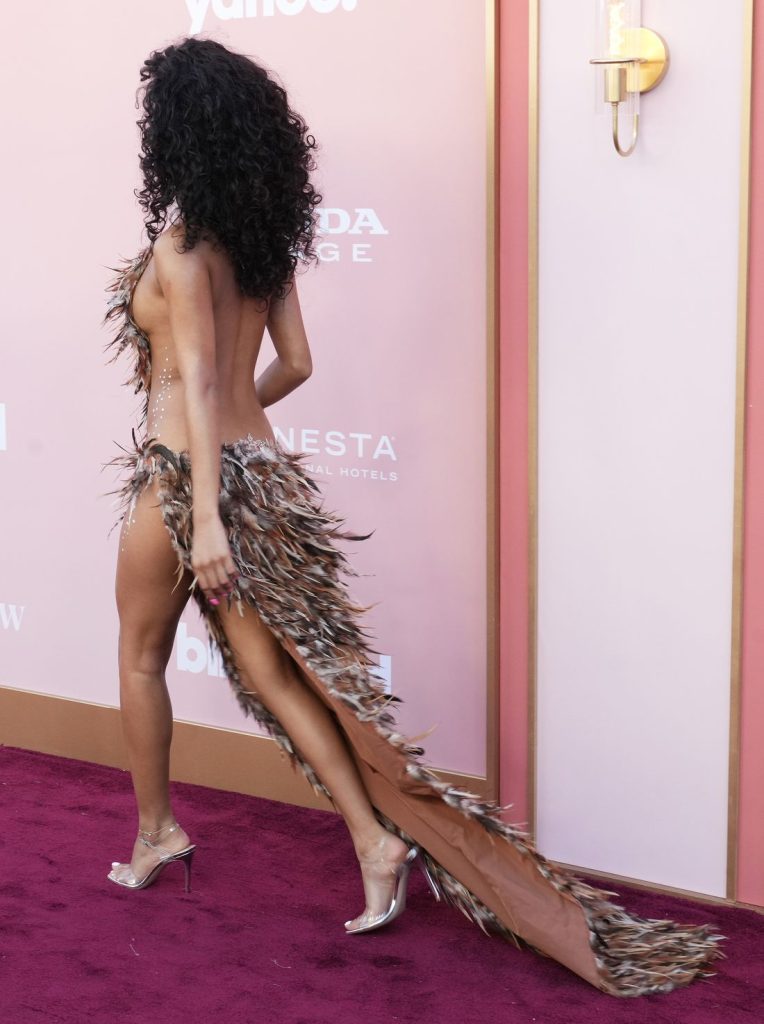 A woman with curly hair walks on a maroon carpet wearing a dramatic gown embellished with feathers, featuring a high slit and train. She is in profile, with her face turned away from the camera.