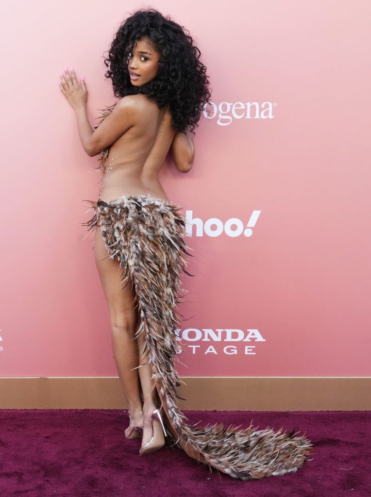 A woman with curly black hair poses on a red carpet in a revealing outfit made of feathers, showing her bare back. She wears clear high heels and stands in front of a peach-colored backdrop with various brand logos #2.