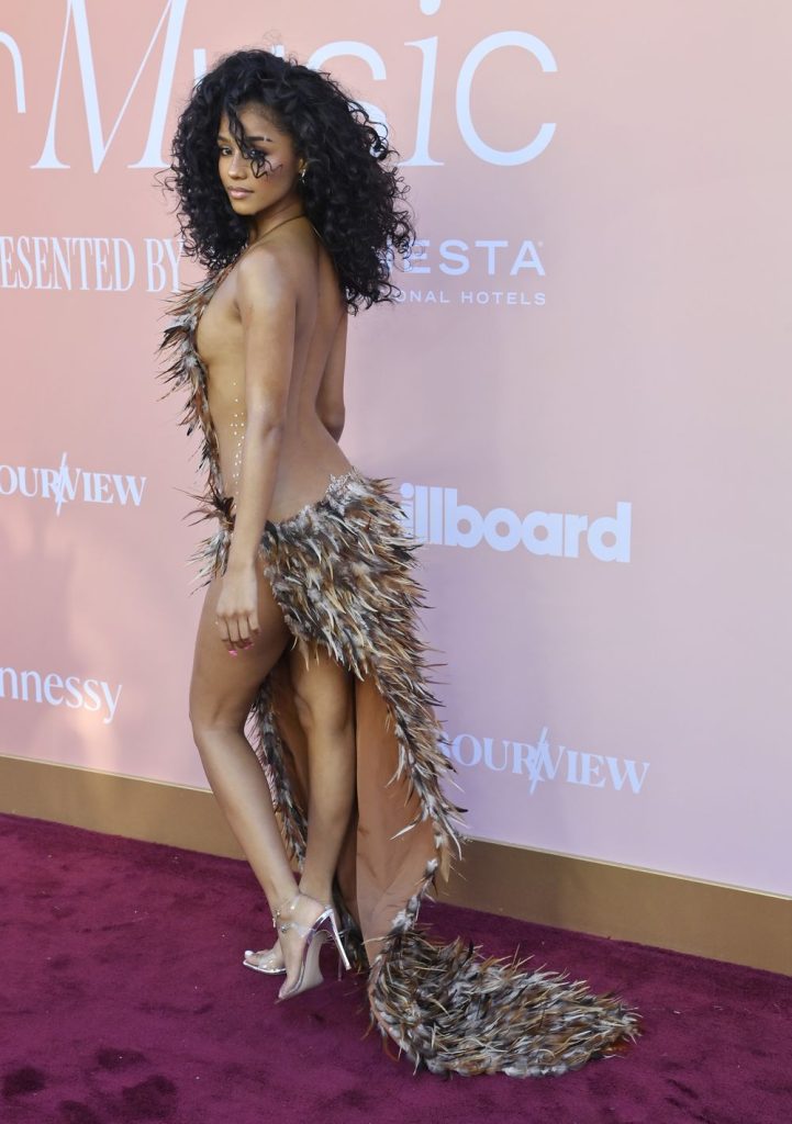 A woman with curly hair poses on a red carpet wearing a bold, feathered outfit and high heels, with dramatic eye makeup. The background displays logos.