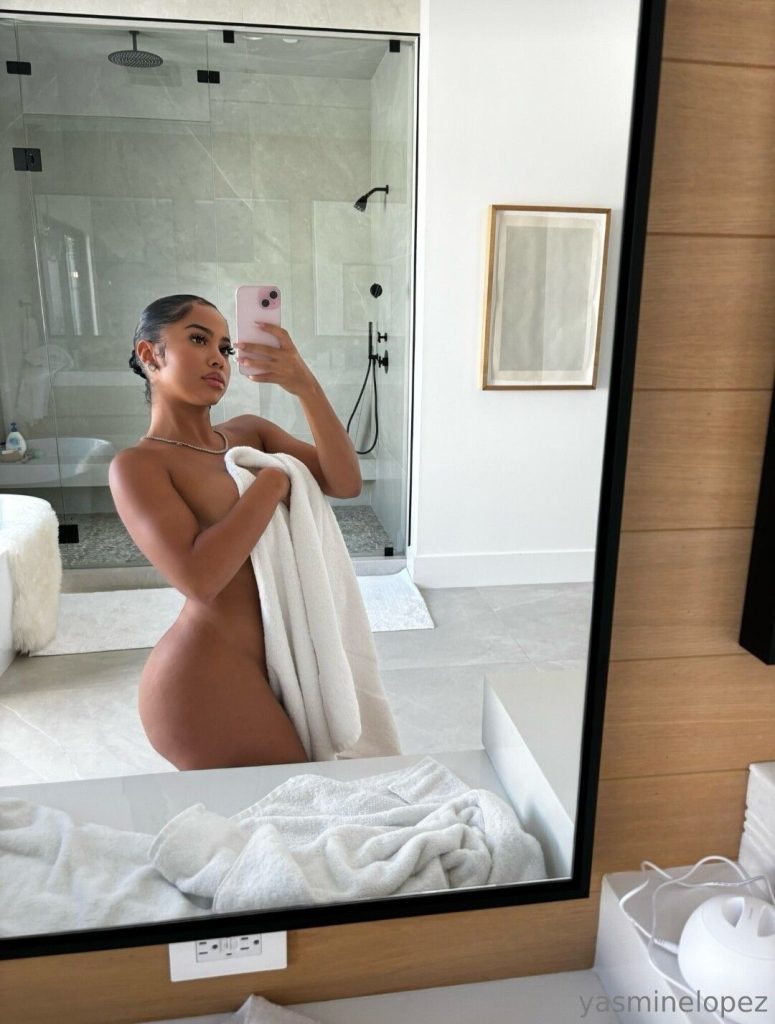 A woman stands in a modern bathroom, holding a white towel to cover herself while taking a mirror selfie with her phone. The background features a glass shower and minimalistic decor.