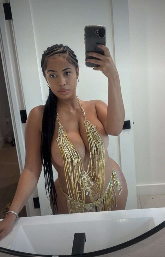 A woman with long braids takes a mirror selfie in a bathroom while wearing a revealing outfit made of gold chains.