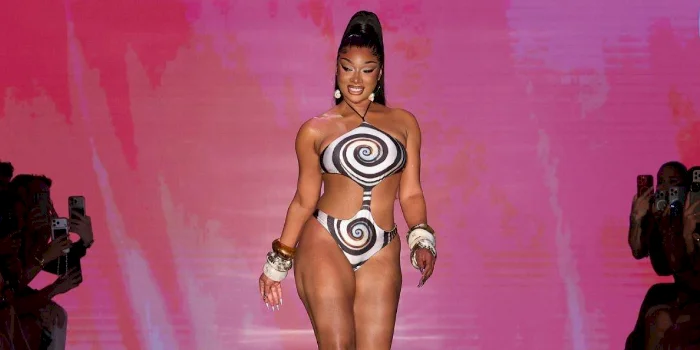 Megan Thee Stallion walks down a runway wearing a bold bikini with black and white swirl patterns, gold accessories, and high heels. The background is pink with an audience seated on both sides.
