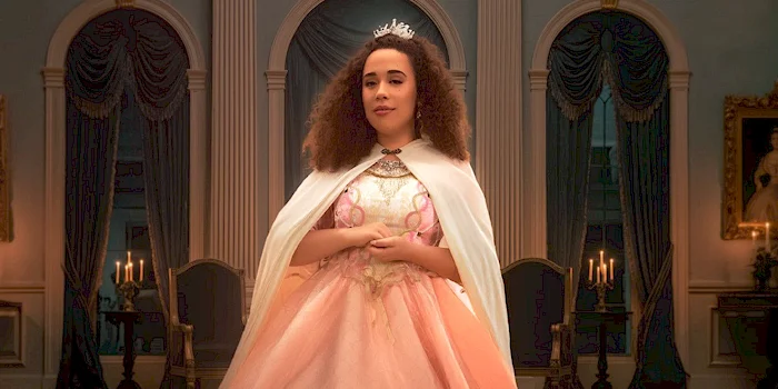 Willow Ryder in a pink ballgown and white cape stands in an elegant, candle-lit room, wearing a tiara.