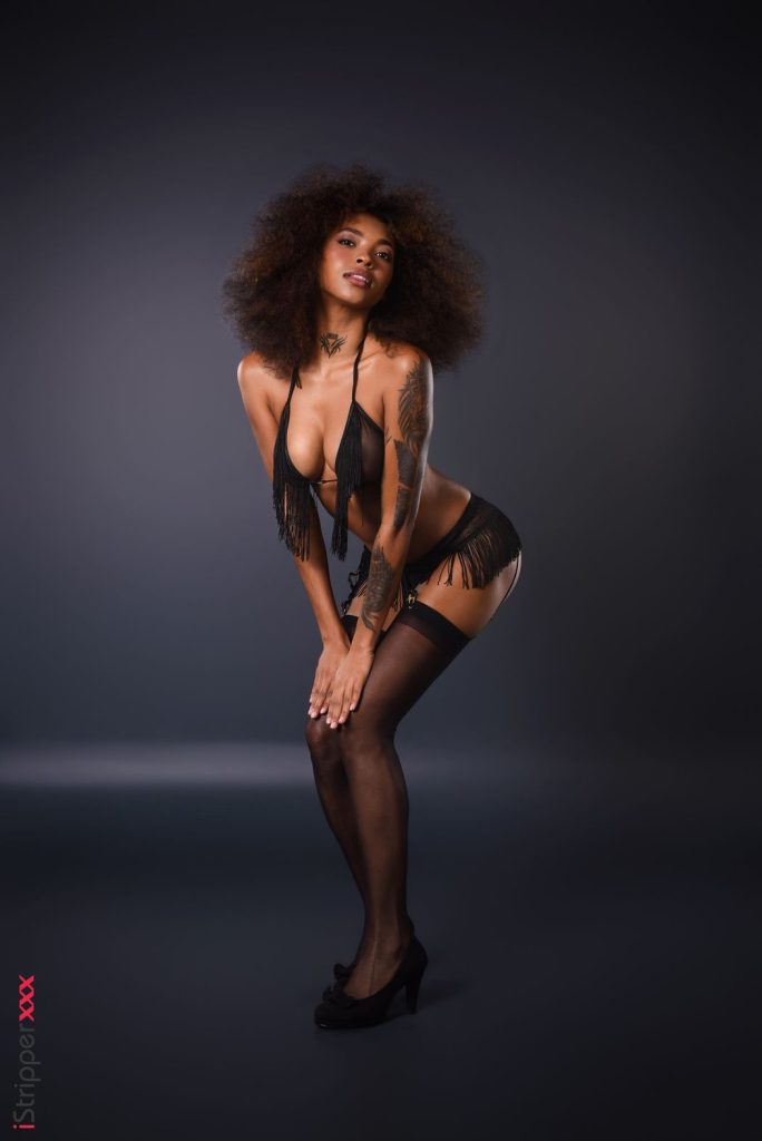 A Latina woman with an afro hairstyle poses in black lingerie, thigh-high stockings, and high heels against a dark studio background. She is slightly bent forward with her hands on her knees, looking confidently at the camera.