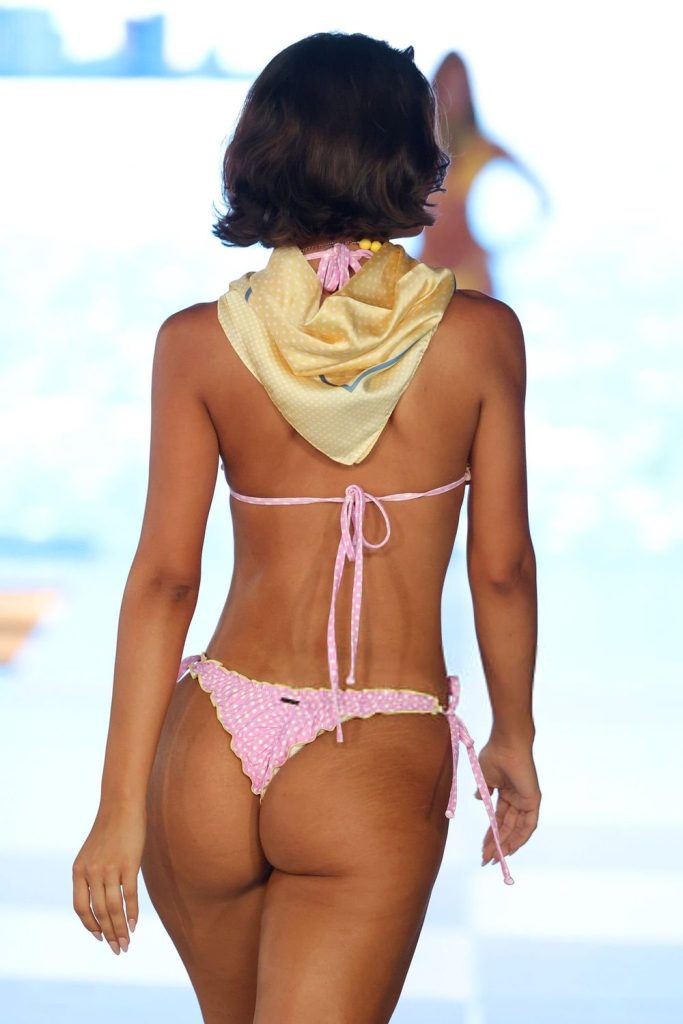 A model walks on a runway wearing a light pink polka dot bikini with frilled edges and a yellow scarf tied around her neck. The background is brightly lit and out of focus.