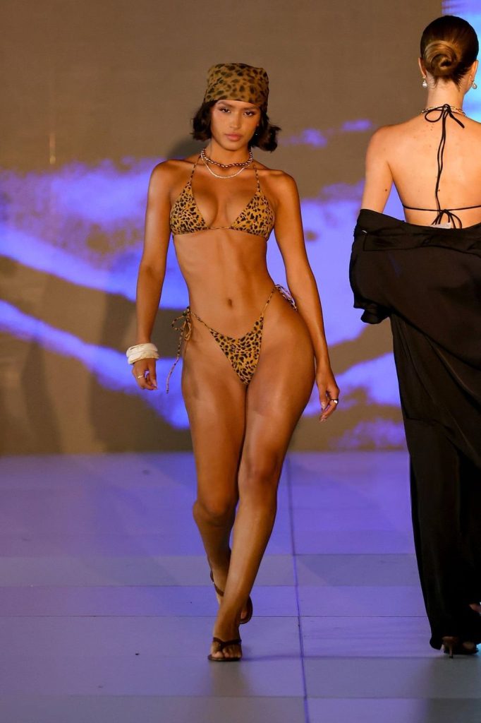 A model walks down a runway wearing a leopard print bikini and matching headscarf, accessorized with a white bracelet, against a blue and purple lit background. Another model in a black outfit walks ahead of her.