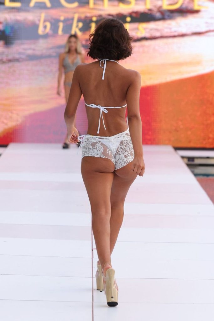 A woman walks down a runway wearing white lace bikini bottoms and a bikini top, with high heels. Another model is visible in the background, and a beach-themed backdrop is displayed behind them.