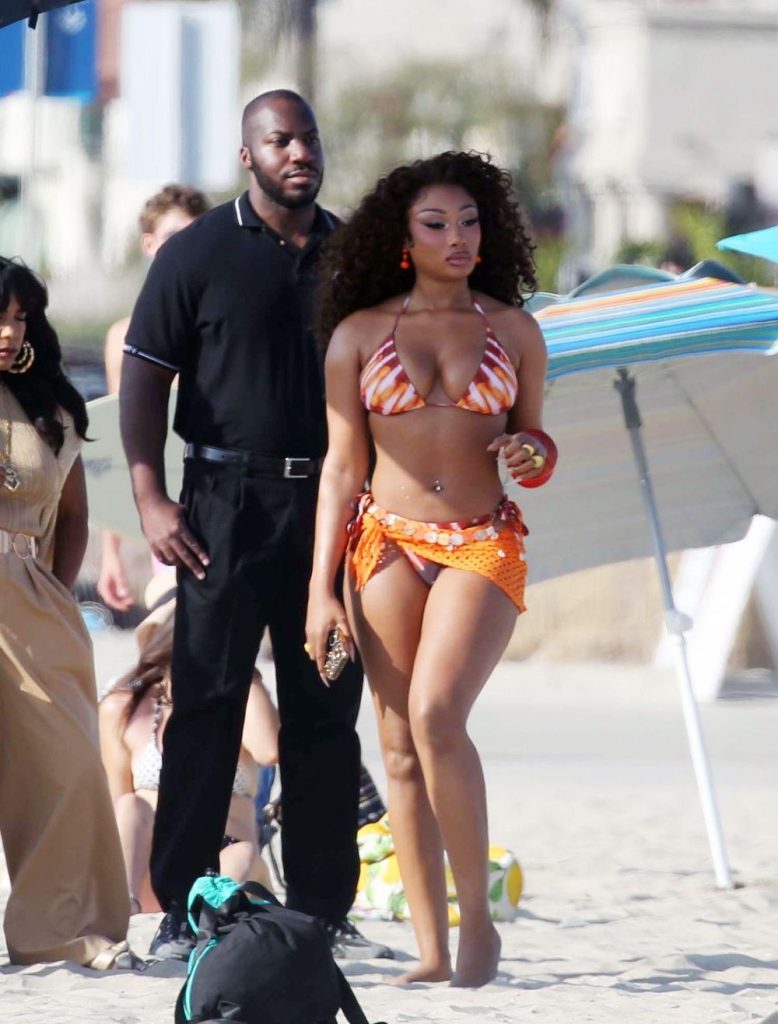 Megan Thee Stallion in an orange and white bikini with a matching sarong walks on a sandy beach, holding a phone and wearing bracelets. A man in a black shirt and pants stands nearby, with people and umbrellas in the background.