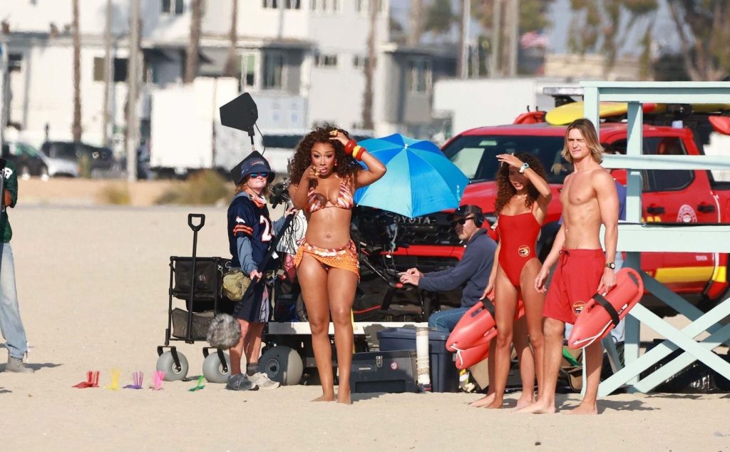 Actors dressed as lifeguards in red swimsuits stand on a beach near a lifeguard stand, with film crew and equipment visible in the background. The scene appears to be part of a film or TV production.