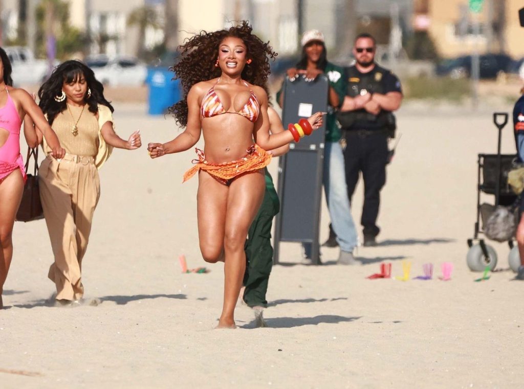 Megan Thee Stallion in an orange bikini runs on the beach, smiling, followed by others in colorful outfits. Police officers and bystanders watch in the background. The scene appears lively and energetic.