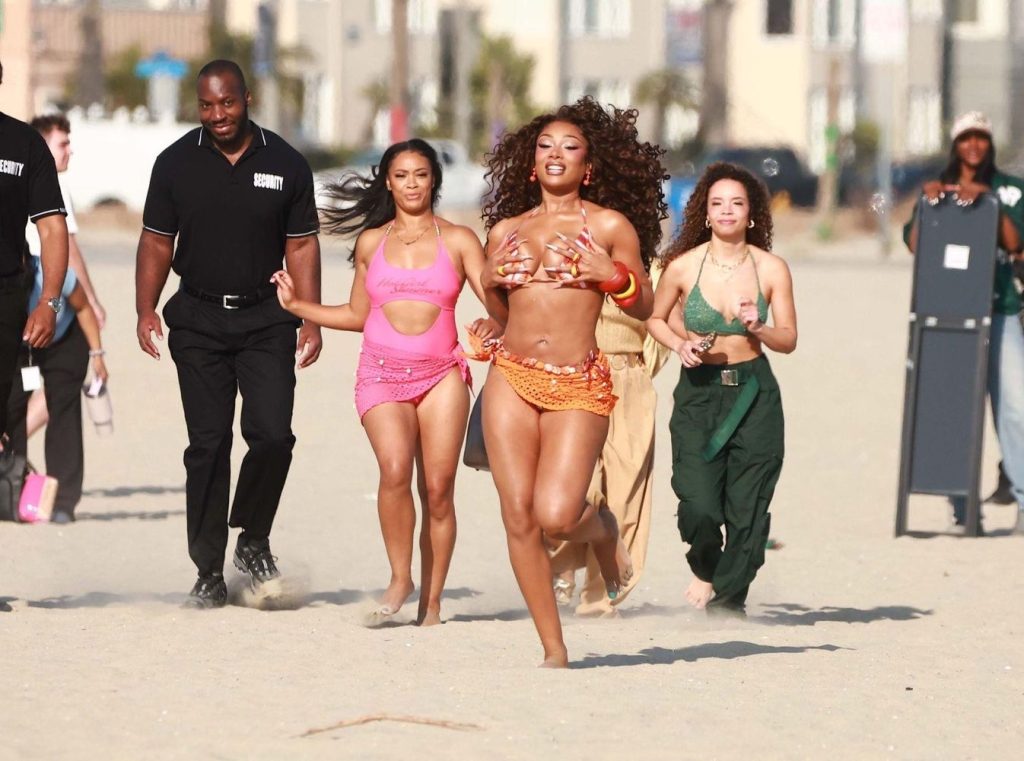 Three women in colorful outfits run joyfully on a sandy beach, followed by security personnel and others. The background shows blurred buildings and palm trees.
