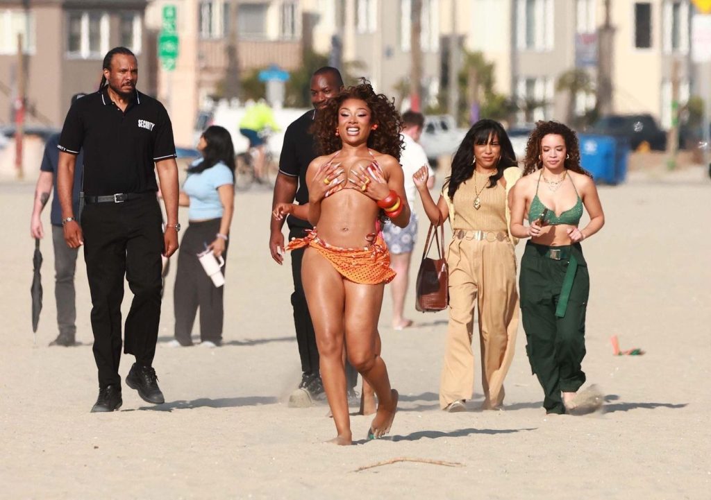 Megan Thee Stallion in a glittery orange outfit runs joyfully on a sandy beach, followed by three women and a security guard. Buildings and other people appear in the background.
