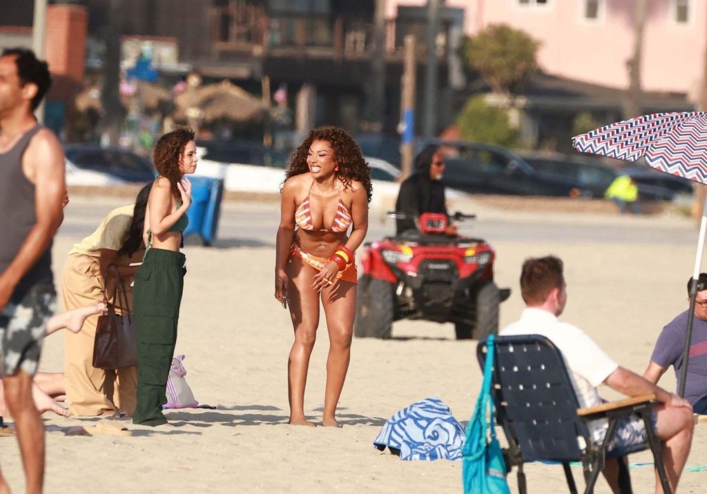 Megan Thee Stallion in a red and white bikini stands smiling on a busy beach surrounded by people, with a red ATV and buildings visible in the background.