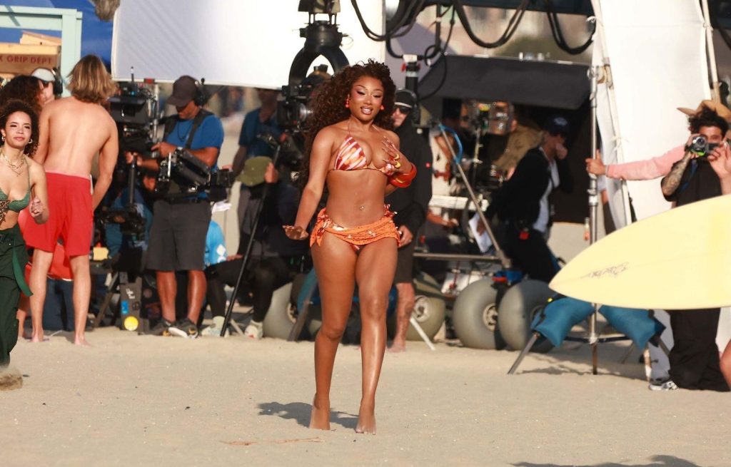 Megan Thee Stallion in an orange bikini smiles while walking on a beach, surrounded by production crew, cameras, and film equipment, with other people and a surfboard nearby.