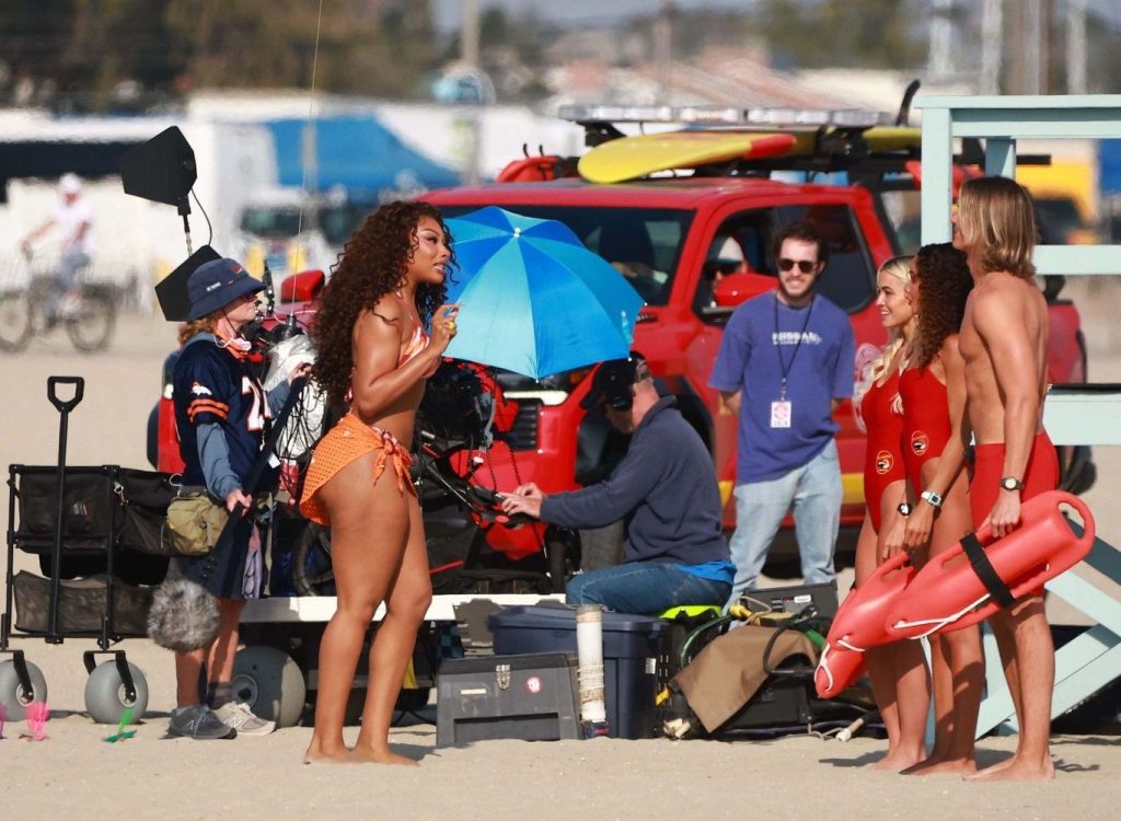 Megan Thee Stallion in an orange swimsuit talks to three lifeguards, two women and one man, on a beach film set with crew members, equipment, and a red vehicle in the background.