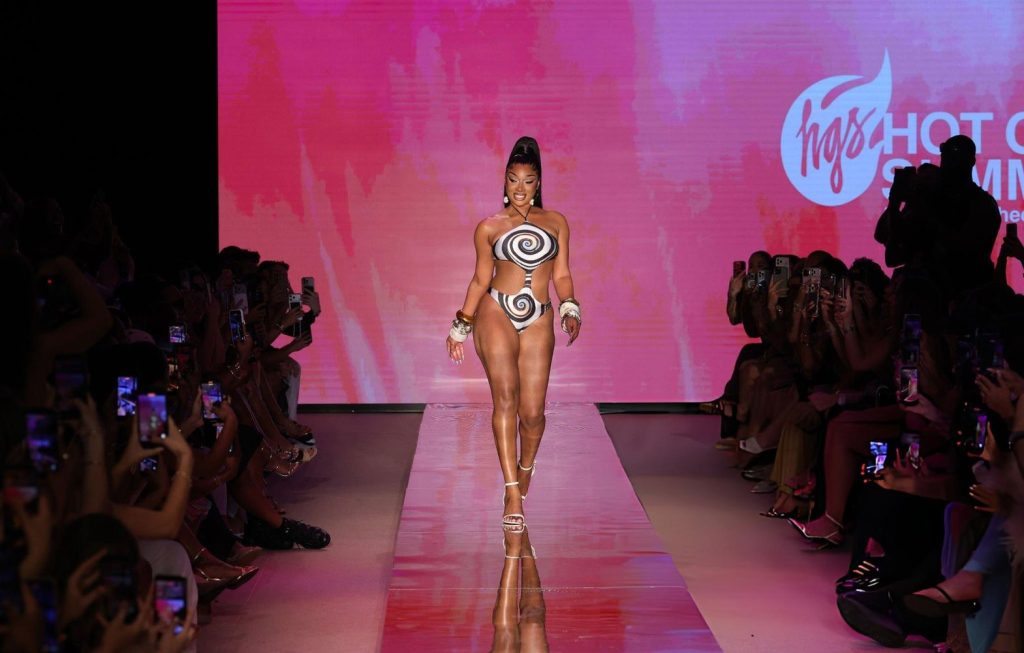 Megan Thee Stallion walks down a runway wearing a bold bikini with black and white swirl patterns, gold accessories, and high heels. The background is pink with an audience seated on both sides and a logo displayed on the screen.