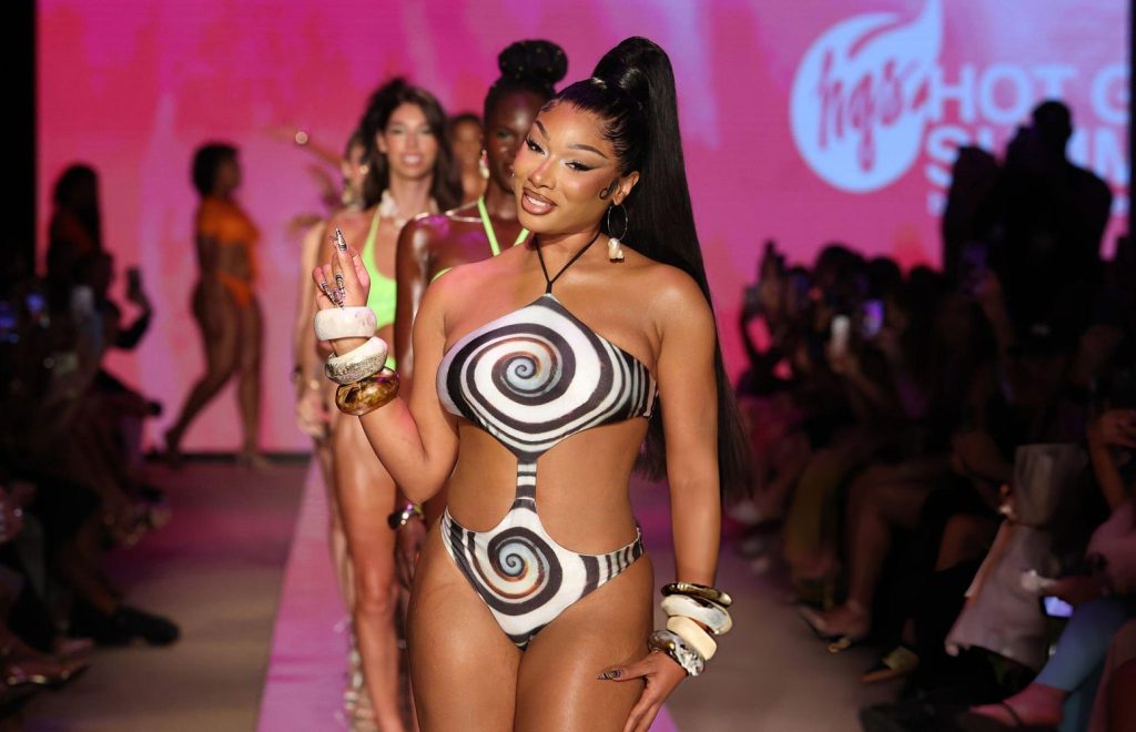 Close up of Megan Thee Stallion stands on a runway wearing a bold, black and white spiral-patterned cutout swimsuit, large bangles, and high heels. Other models and an audience are visible, with a pink-lit background and a show logo.