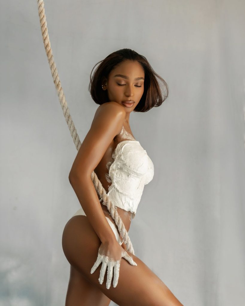 A woman with short dark hair poses sideways on a thick rope, wearing white textured fabric wrapped around her chest and paint on her hand, against a soft gray background.
