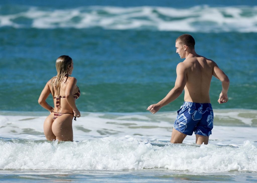 A woman in a bikini and a man in blue swim trunks are wading into the ocean, with gentle waves and blue water in the background.