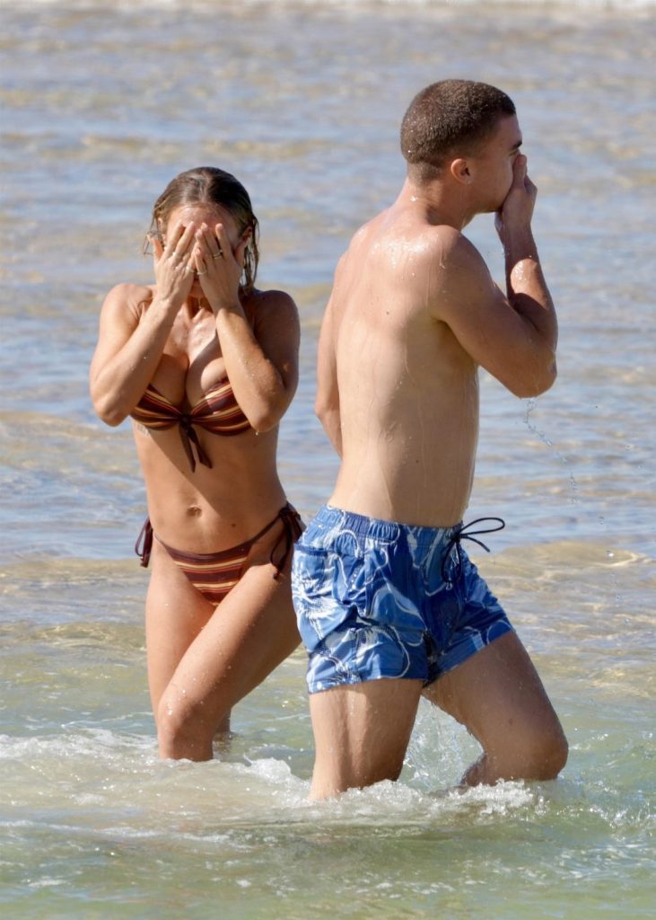 A woman in a brown bikini and a man in blue swim shorts walk out of the ocean, both covering their faces with their hands as water drips from them. The background shows shallow, calm sea water.