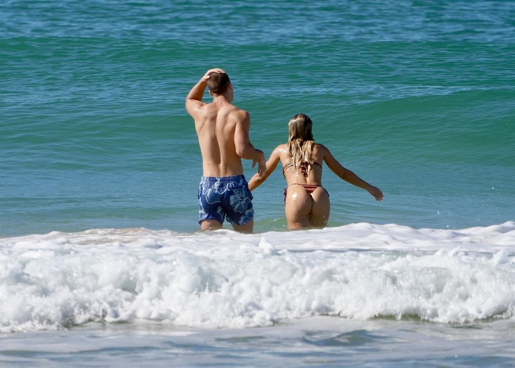 A man and a woman in swimwear are holding hands and walking into the ocean together, with waves around them on a sunny day.