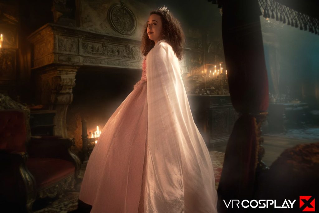 A woman in a pale pink gown and white cape, wearing a tiara, stands in an ornate, dimly lit room with a grand fireplace and candles.