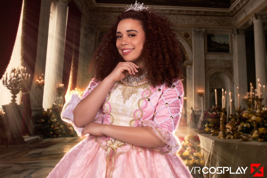 A woman with curly hair wears a pink, ornate princess gown and a tiara, smiling confidently in an elegant, lavishly decorated hall with chandeliers and banquet tables.