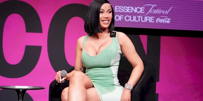 Woman in a green and white dress sits onstage with a microphone, smiling. Behind her is a pink backdrop and a sign that reads Essence Festival of Culture.