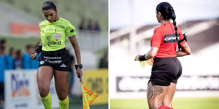 Two female soccer referees are pictured on a field, one in a yellow shirt holding a flag, and the other in a red shirt with a visible leg tattoo. Both are focused on their tasks during a match.