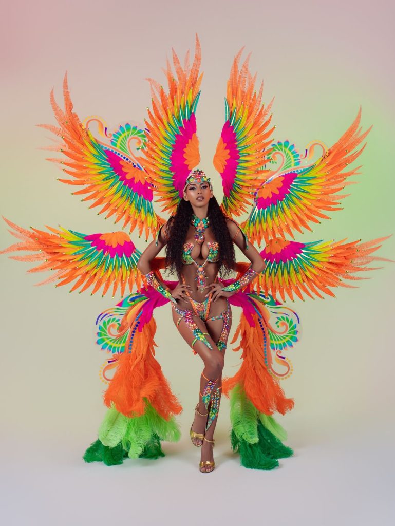 A woman poses in a vibrant carnival costume with orange, green, and pink feathers, jeweled accessories, and large wing-like structures, standing against a gradient pastel background.