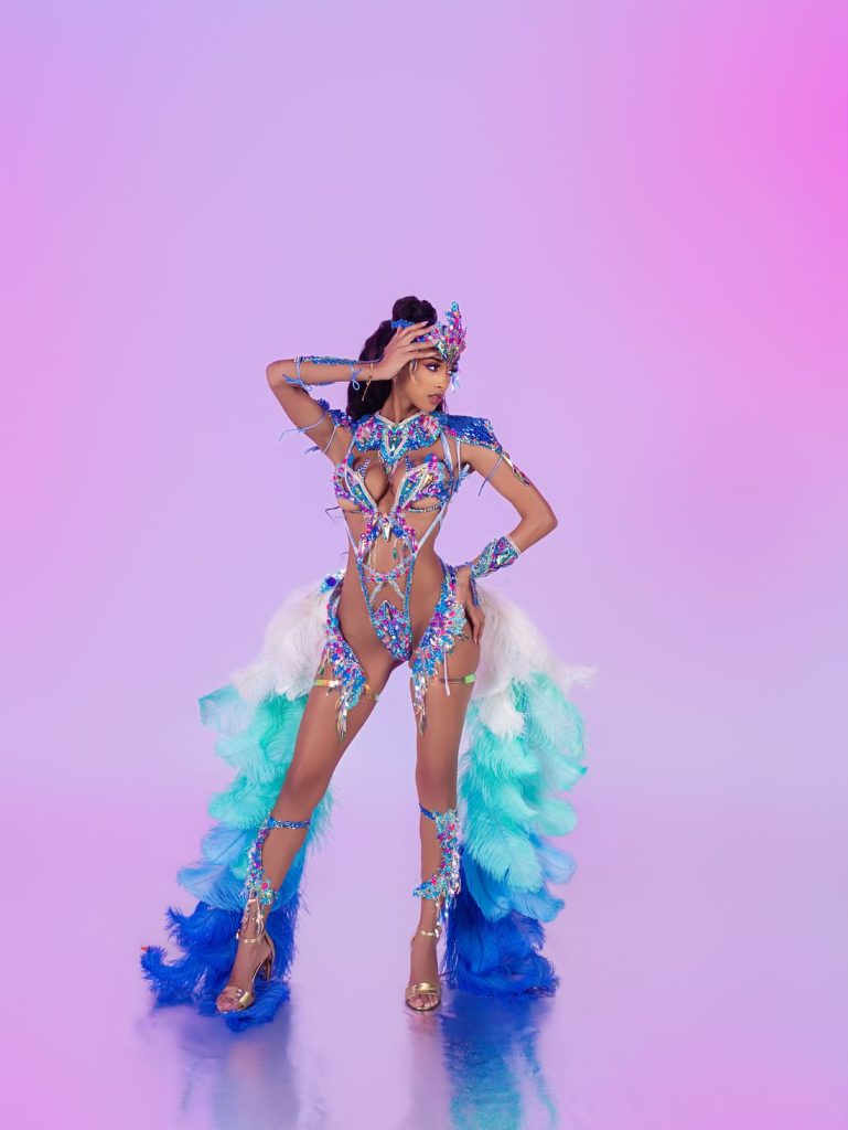 A woman poses in a colorful, feathered carnival costume with blue, turquoise, and white accents, sparkling jewels, and high heels, standing against a pink to purple gradient background.