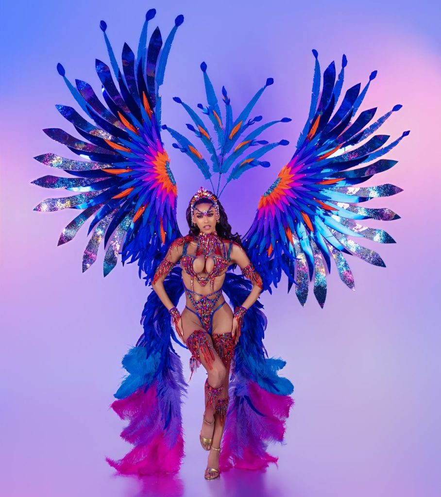 A woman poses in a vibrant, feathered carnival costume with large blue and purple wings, beaded accessories, and a matching headpiece, against a colorful pastel background.