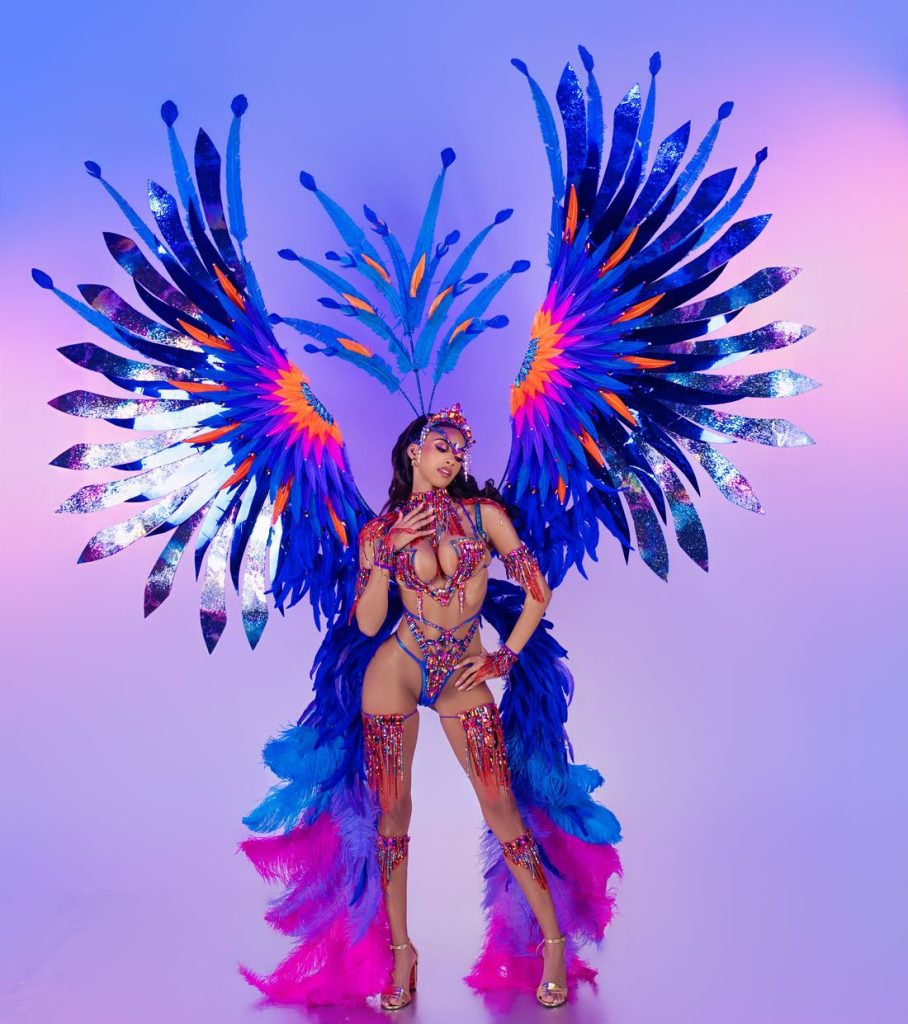 A woman poses in a vibrant carnival costume with large, blue and purple feathered wings, beaded accents, and a matching headdress, set against a gradient pink and purple background.