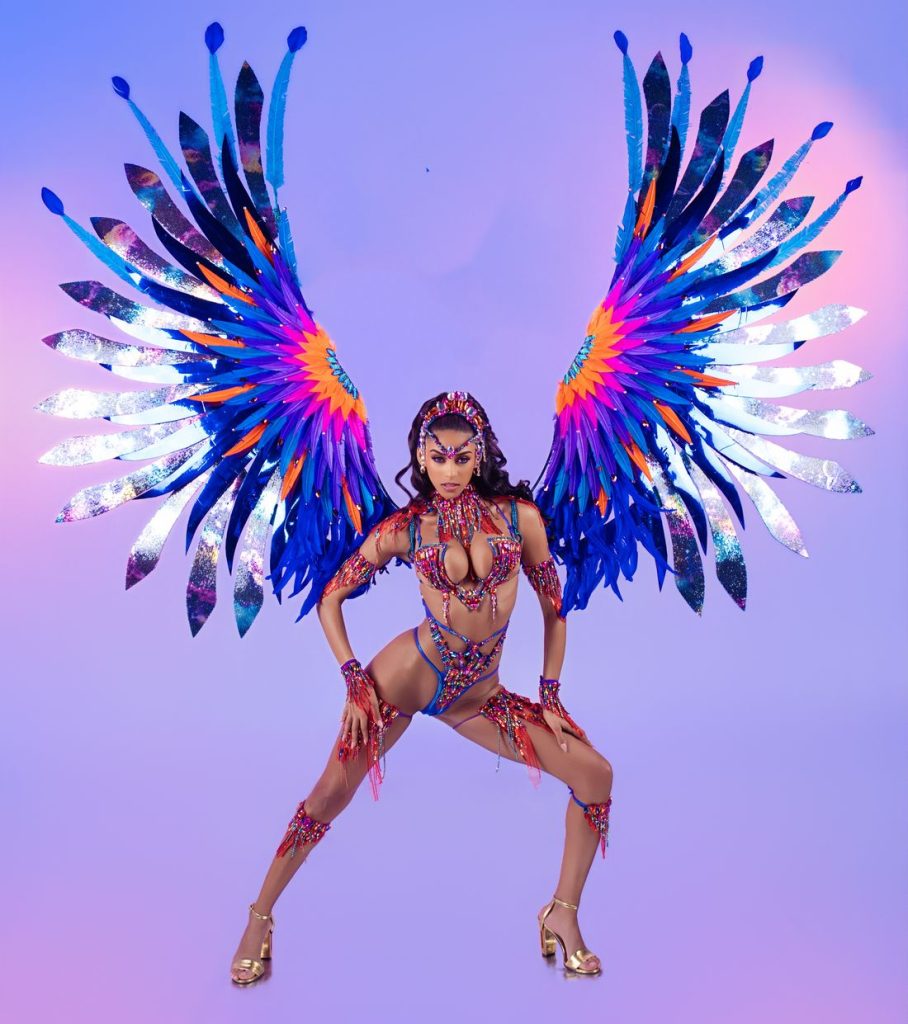 A woman poses in a vibrant carnival costume with large, colorful blue, purple, orange, and pink feathered wings against a gradient pink and blue background.