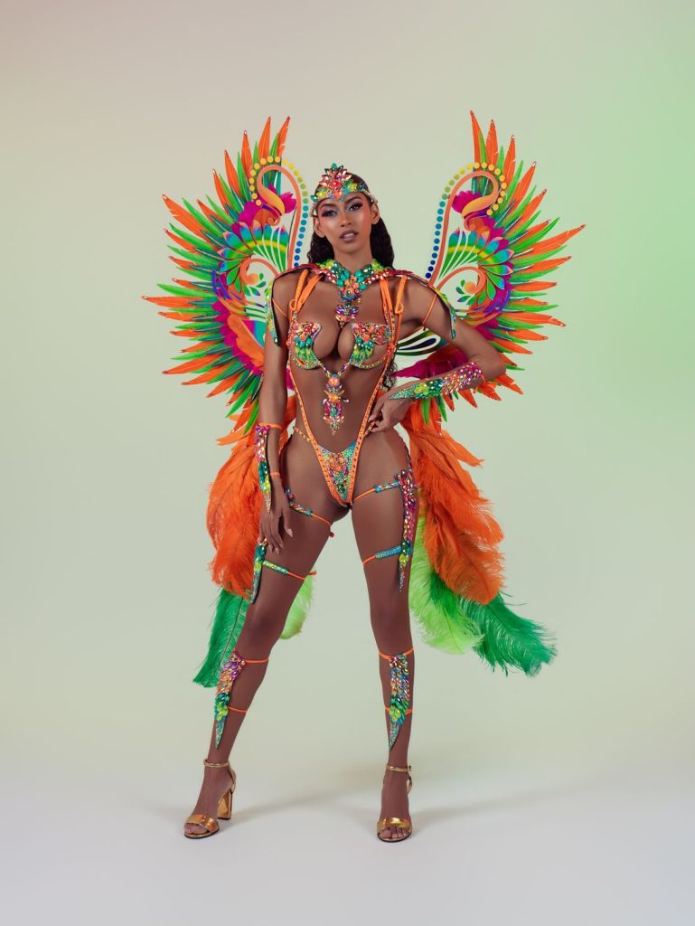 A woman poses in a vibrant Carnival costume with feathered wings in orange, green, and yellow, jeweled accents, and gold heels, standing against a light background.