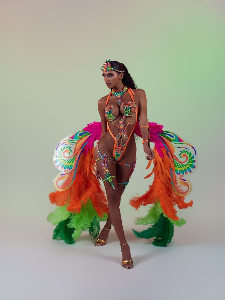 A woman poses in a vibrant carnival costume with neon colors, large feathered wings, beaded details, and gold heels on a light gradient background.