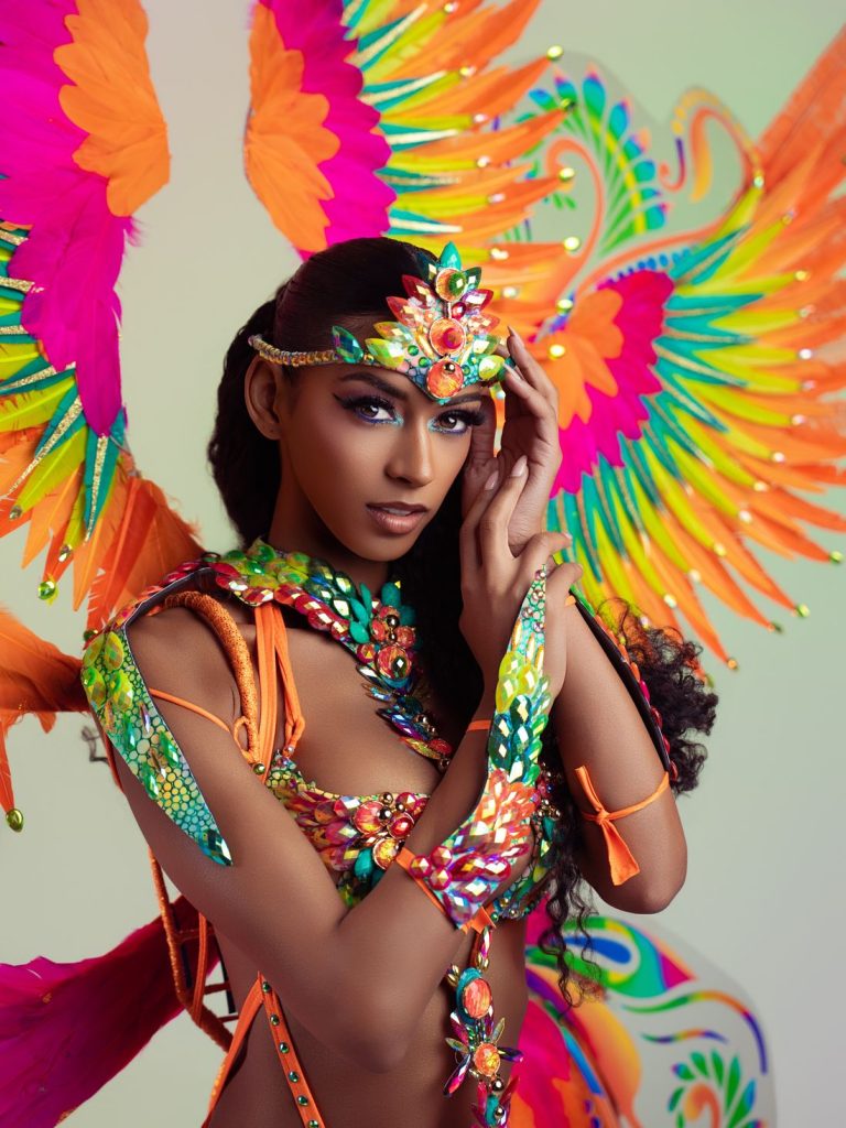 A woman in a vibrant, colorful carnival costume poses with jeweled accessories, orange and green feathered wings, and intricate headpiece, exuding confidence and festive energy.