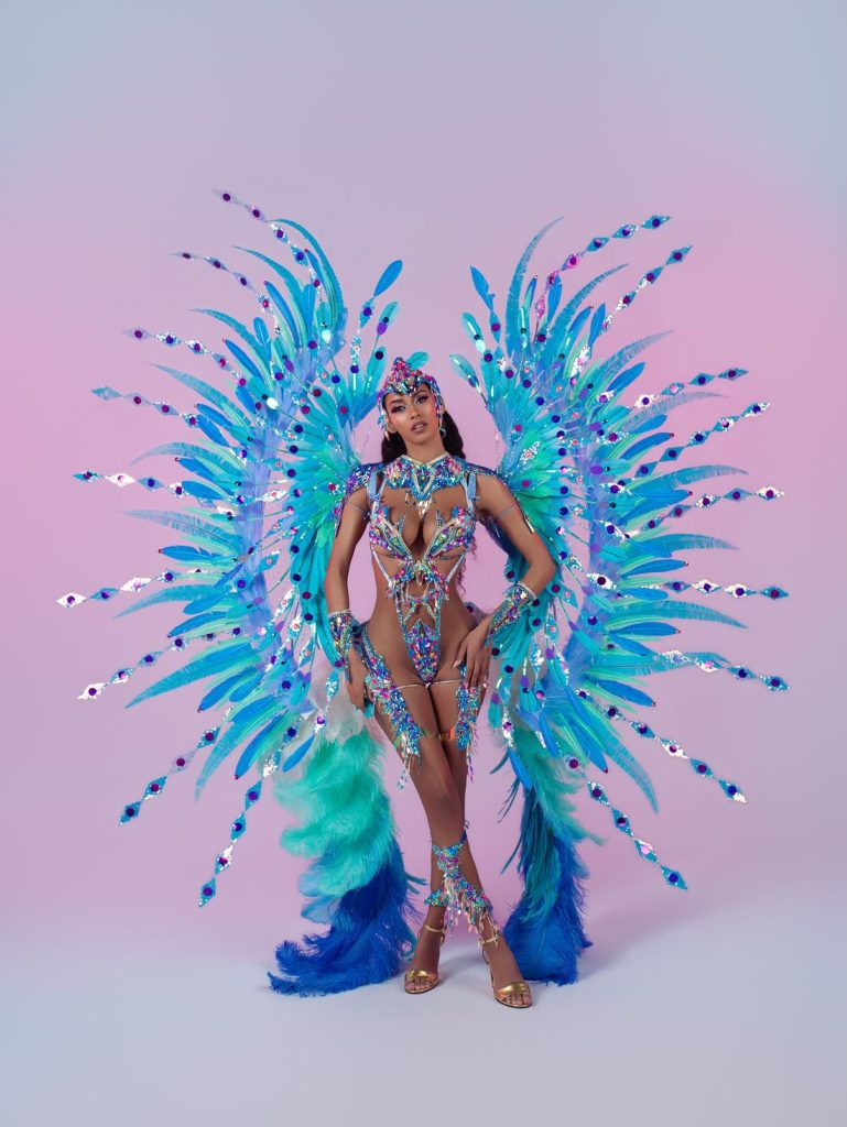A woman poses in a vibrant carnival costume with blue and purple feathers, jeweled details, and large decorative wings against a pink and blue gradient background.