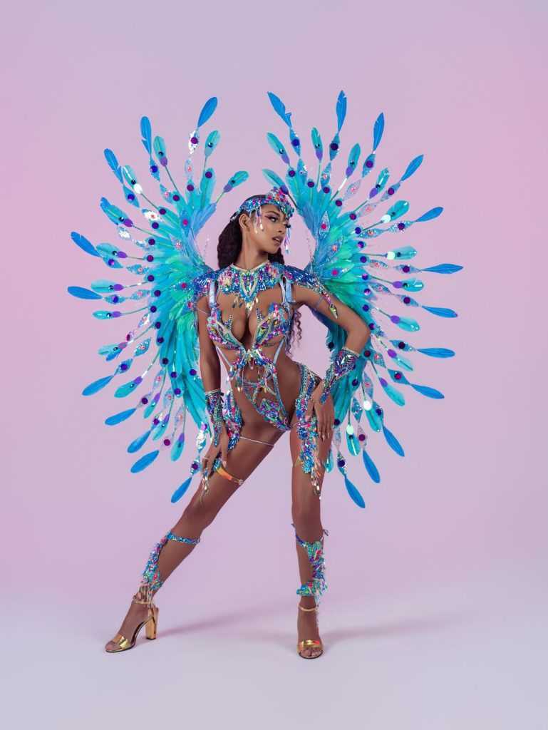 A woman poses in a vibrant, jeweled carnival costume with blue and purple feathers forming dramatic wings, matching headpiece, gold heels, and intricate body adornments, set against a light pink background.