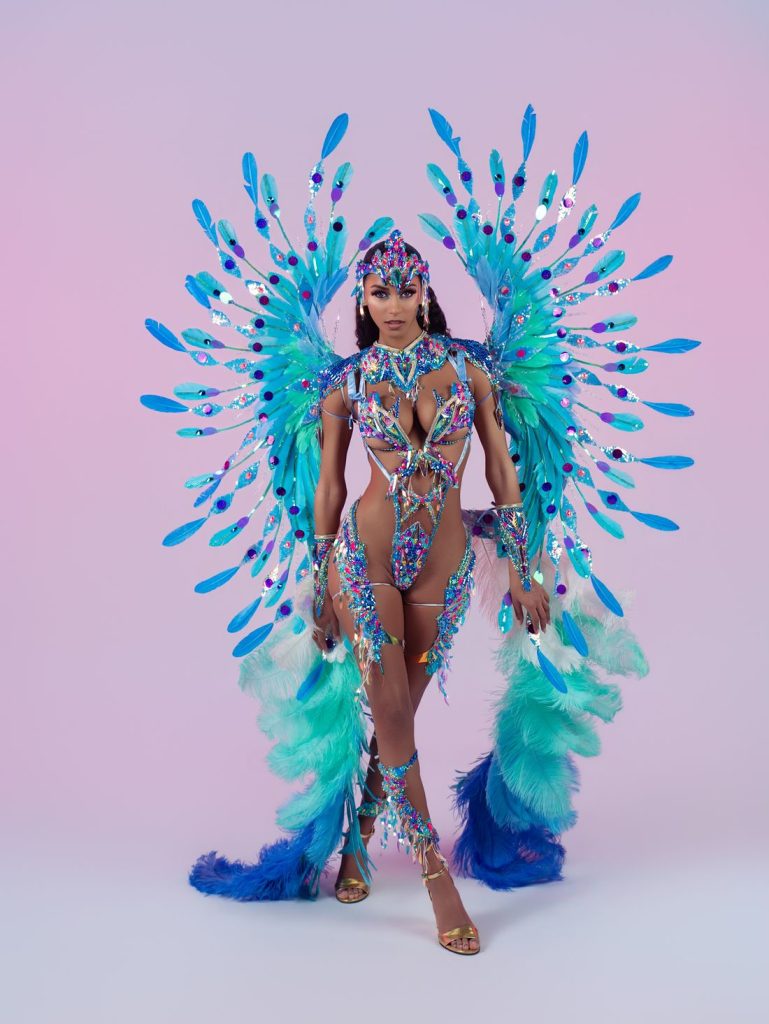 A person wearing a vibrant, feathered carnival costume in blue, turquoise, and purple stands against a pink and purple gradient background. The costume features dramatic wing-like feathers and sparkling embellishments.