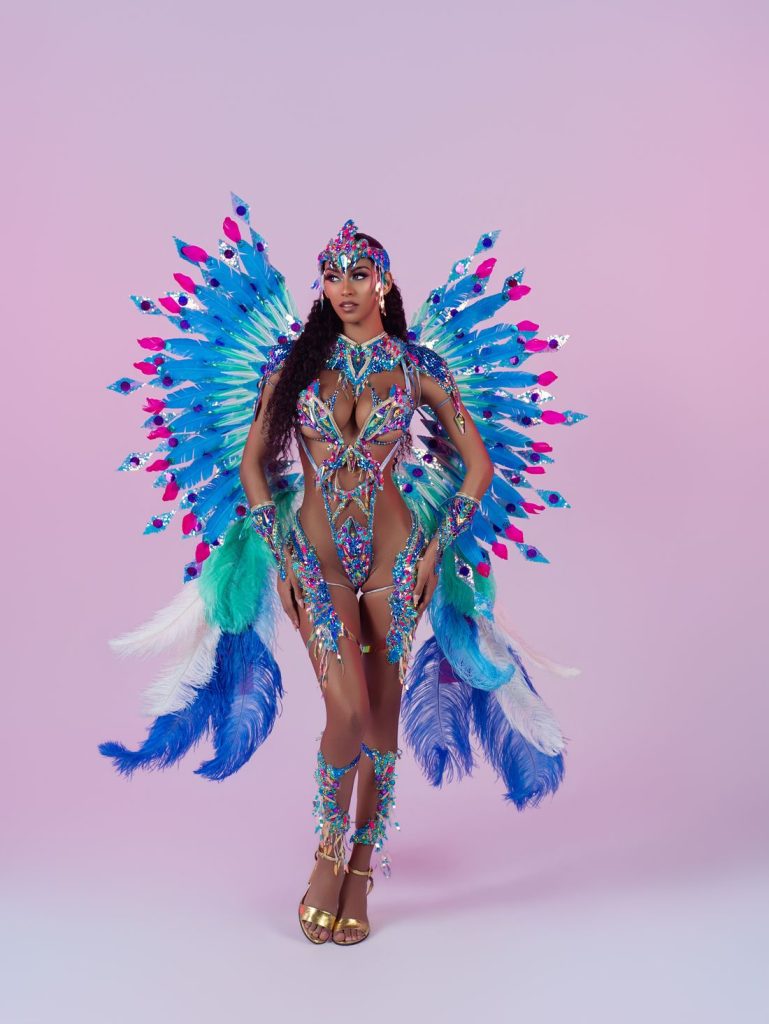 A woman in a vibrant blue, purple, and green feathered carnival costume with intricate beadwork, large feathered wings, and a jeweled headpiece stands confidently against a pink background.