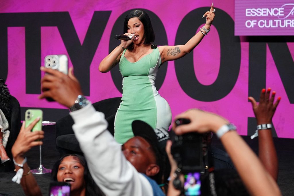 A woman in a green dress sings on stage with a microphone as fans in the audience take photos and videos with their phones. A pink backdrop displays the Essence Festival of Culture logo.