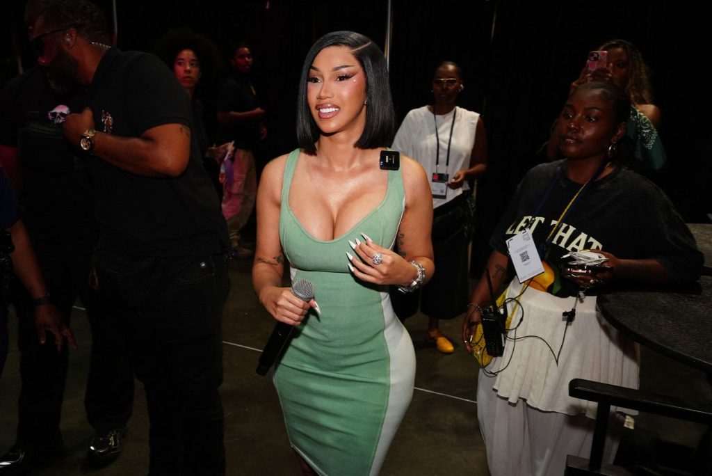 A woman in a green dress holds a microphone and smiles while walking indoors, surrounded by people. Some wear badges and hold equipment. The setting appears to be backstage at an event.