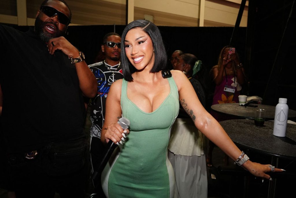 Woman in a green dress smiles and holds a microphone while standing near a group of people. A man in sunglasses stands beside her. A water bottle and drinks are on a tall table nearby.