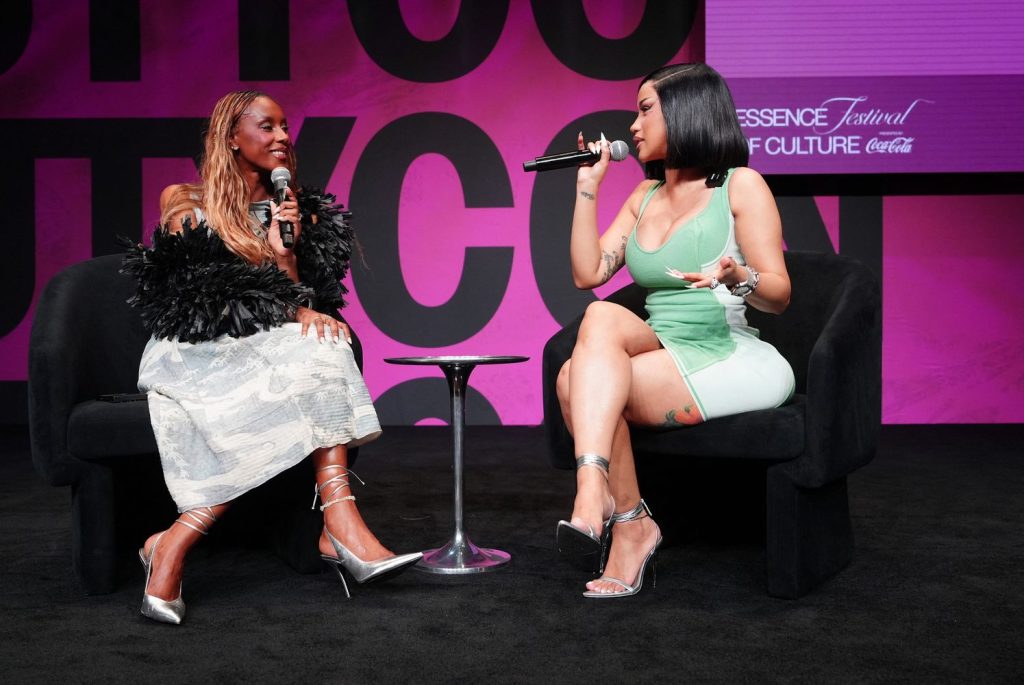 Two women sit on black chairs onstage, holding microphones and talking. The background is pink with text, and a “ESSENCE Festival of Culture” sign is visible. They are dressed stylishly and appear to be in conversation.